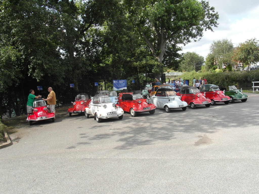 A line of microcars parked diagonally at an outdoor gathering, with a banner and mature trees behind