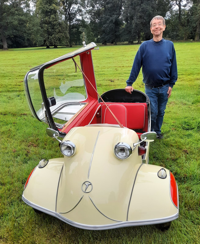 A man standing beside a cream and red Messerschmitt KR200 with its canopy hinged open, on a lawn beneath mature trees
